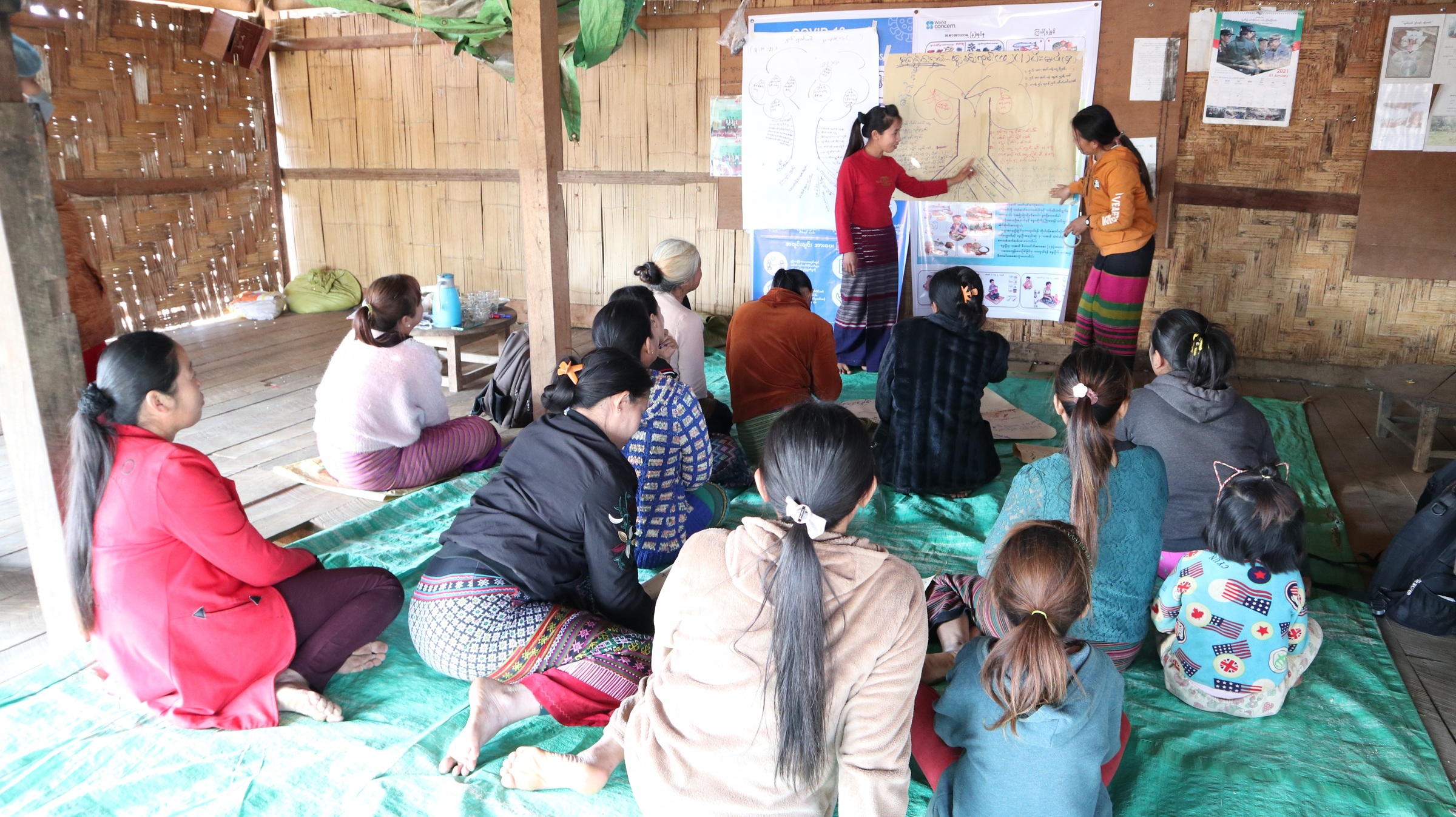 Frauen und Kinder sitzen auf dem Boden vor einer Tafel, an der zwei Frauen etwas erklären (Quelle: Kindernothilfepartner)