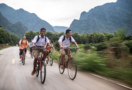 Eine Gruppe jugendlicher Radfahrerinnen und Radfahrer fährt zügig eine Straße entlang, im Hintergrund Dschungel und hohe Berge (Quelle: Jakob Studnar) Eine Gruppe jugendlicher Radfahrerinnen und Radfahrer fährt zügig eine Straße entlang, im Hintergrund Dschungel und hohe Berge (Quelle: Jakob Studnar)